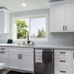 Jasna kuchnia z białymi szafkami z czarnymi uchwytami, szarym blatem i oknem z widokiem na zieleń, nad zmywarką wiszą dwie ściereczki w kratę.