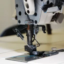 Zbliżenie na stopkę przemysłowej maszyny do szycia z igłą i nitką, widoczne metalowe elementy mechanizmu oraz fragment blatu stołu, oświetlenie studyjne.