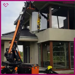 Montaż dużej szyby okiennej przy użyciu specjalistycznego dźwigu z przyssawkami, widoczny pracownik w odblaskowej kamizelce i fragment budynku z ciemną dachówką.