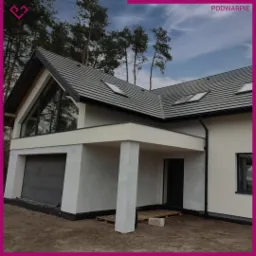 Nowoczesny dom jednorodzinny z szarą dachówką i dużym oknem trójkątnym nad garażem, widoczne elementy wykończeniowe elewacji, częściowo niezagospodarowany teren wokół budynku.