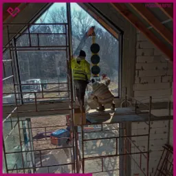 Montaż okna w surowym stanie budynku: dwóch monterów na rusztowaniu, jeden z przyssawkami do szkła, widoczny fragment lasu w tle.