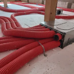 Czerwone, karbowane rury wentylacyjne połączone z metalową skrzynką rozdzielczą na poddaszu z drewnianą konstrukcją dachu, widoczne mocowania rur.