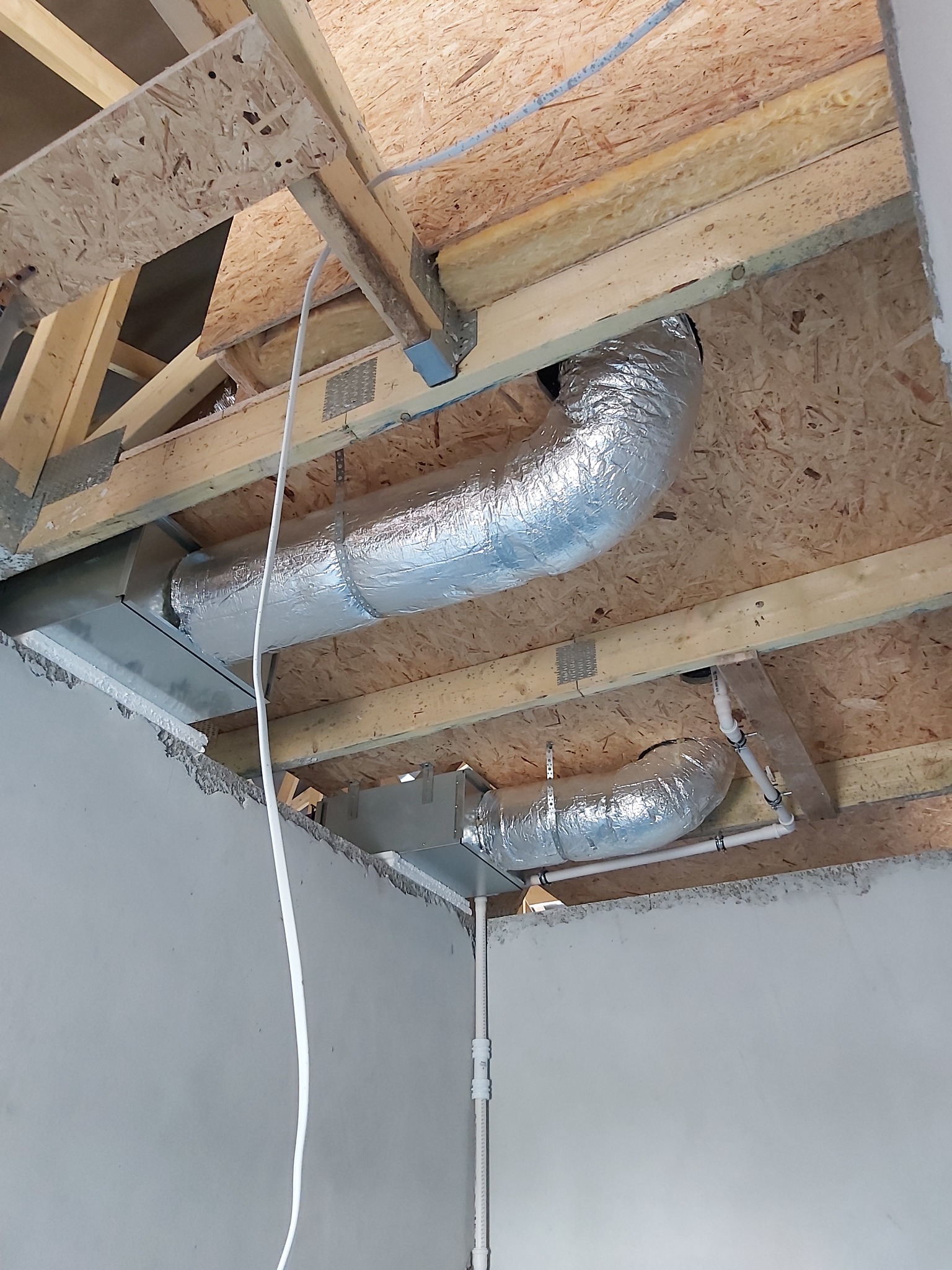 Fragment instalacji z widocznymi kanałami wentylacyjnymi w izolacji aluminiowej, zamontowanymi pod drewnianą konstrukcją stropu na surowym etapie wykończenia budynku.