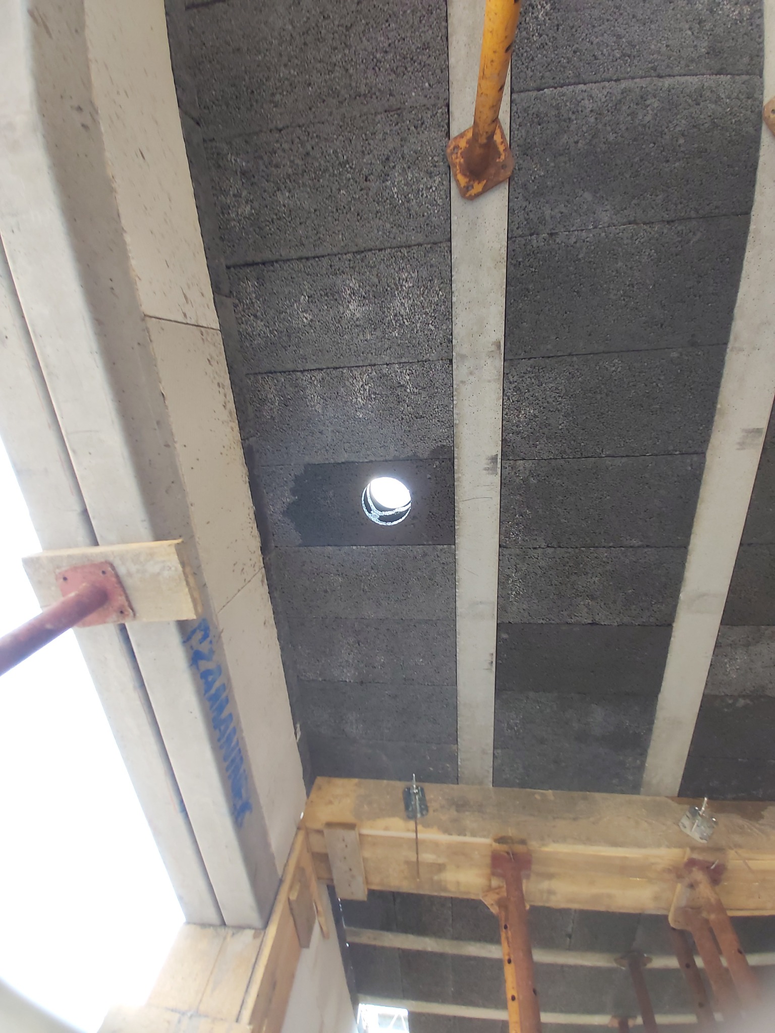 Widok z dołu na strop z pustaków szalunkowych z wywierconym otworem na instalację, wsparty na drewnianych podporach i metalowych stemplach budowlanych.