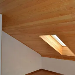 Wnętrze poddasza z drewnianą boazerią na skosach i suficie, okno dachowe wpuszczone w sufit, pomalowana ściana w tle.