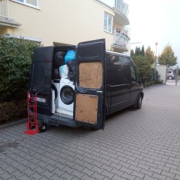 Przeprowadzki Jacek Jasiński - Czarny van z otwartymi drzwiami bagażnika, załadowany pralką, materacem i workami z rzeczami, przed budynkiem mieszkalnym; czerwony wózek transportowy z tyłu pojazdu.
