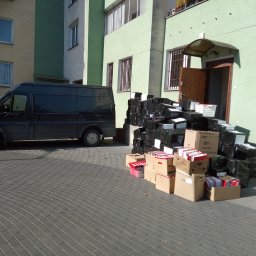 Przeprowadzki Jacek Jasiński - Czarny van dostawczy zaparkowany obok sterty zapakowanych kartonów i folii przed wejściem do budynku.