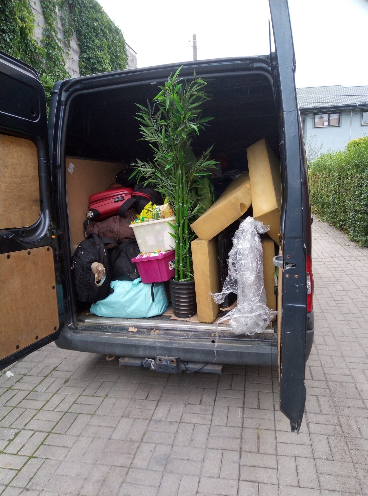 Otwarty bagażnik busa załadowany bagażami, kartonami i sztuczną rośliną, widok z tyłu na kostkę brukową i fragment budynku z zielonym pnączem.