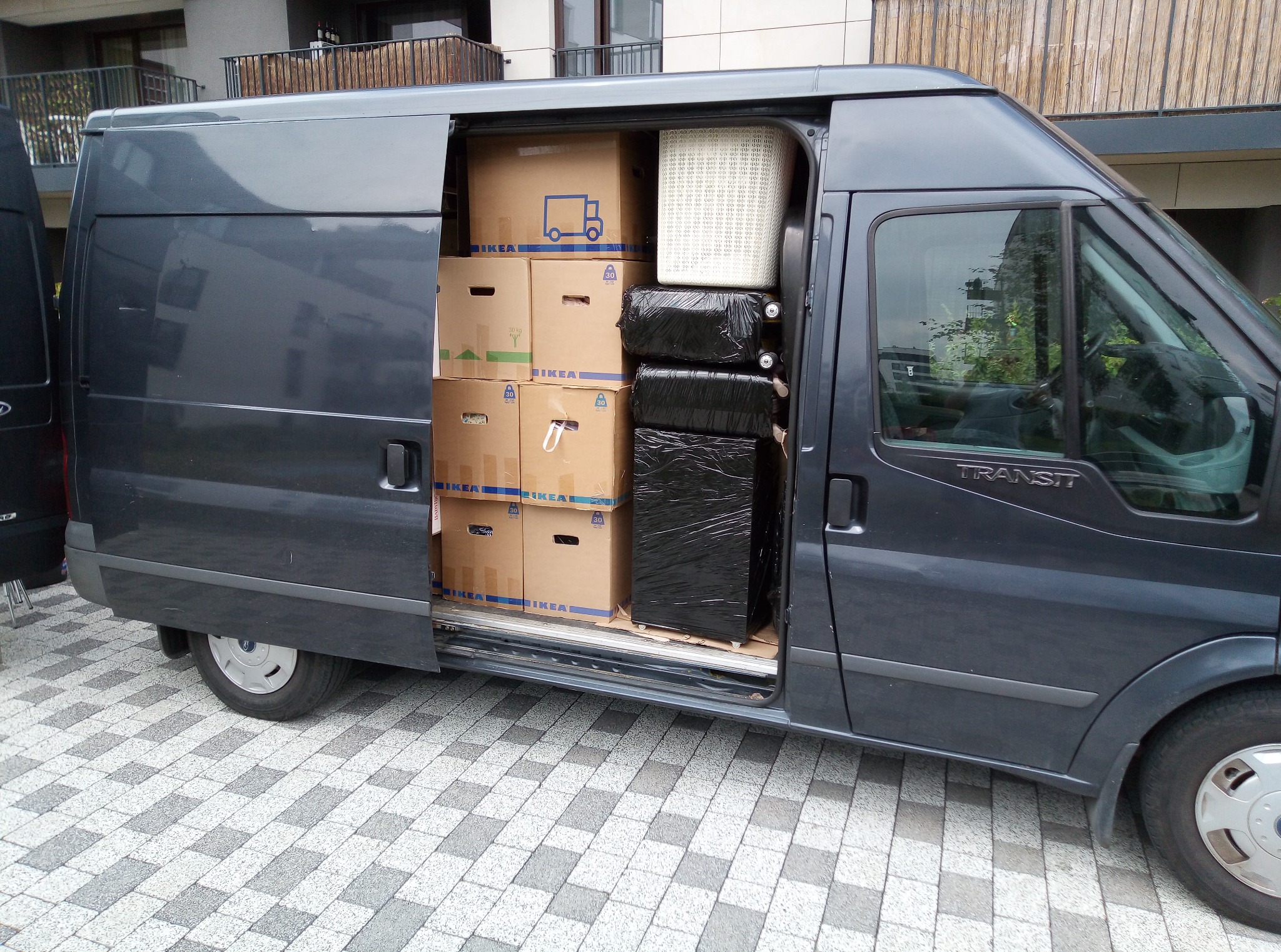 Szary bus marki Ford Transit z otwartymi drzwiami bocznymi, wypełniony kartonami IKEA i bagażami przygotowanymi do przewozu, na tle bloku mieszkalnego.