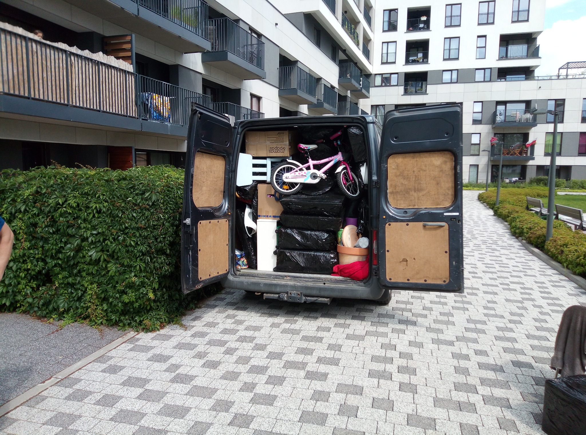 Otwarty bagażnik busa załadowany kartonami, workami i różowym rowerkiem dziecięcym na tle nowoczesnego budynku mieszkalnego z balkonami i brukowanego chodnika.