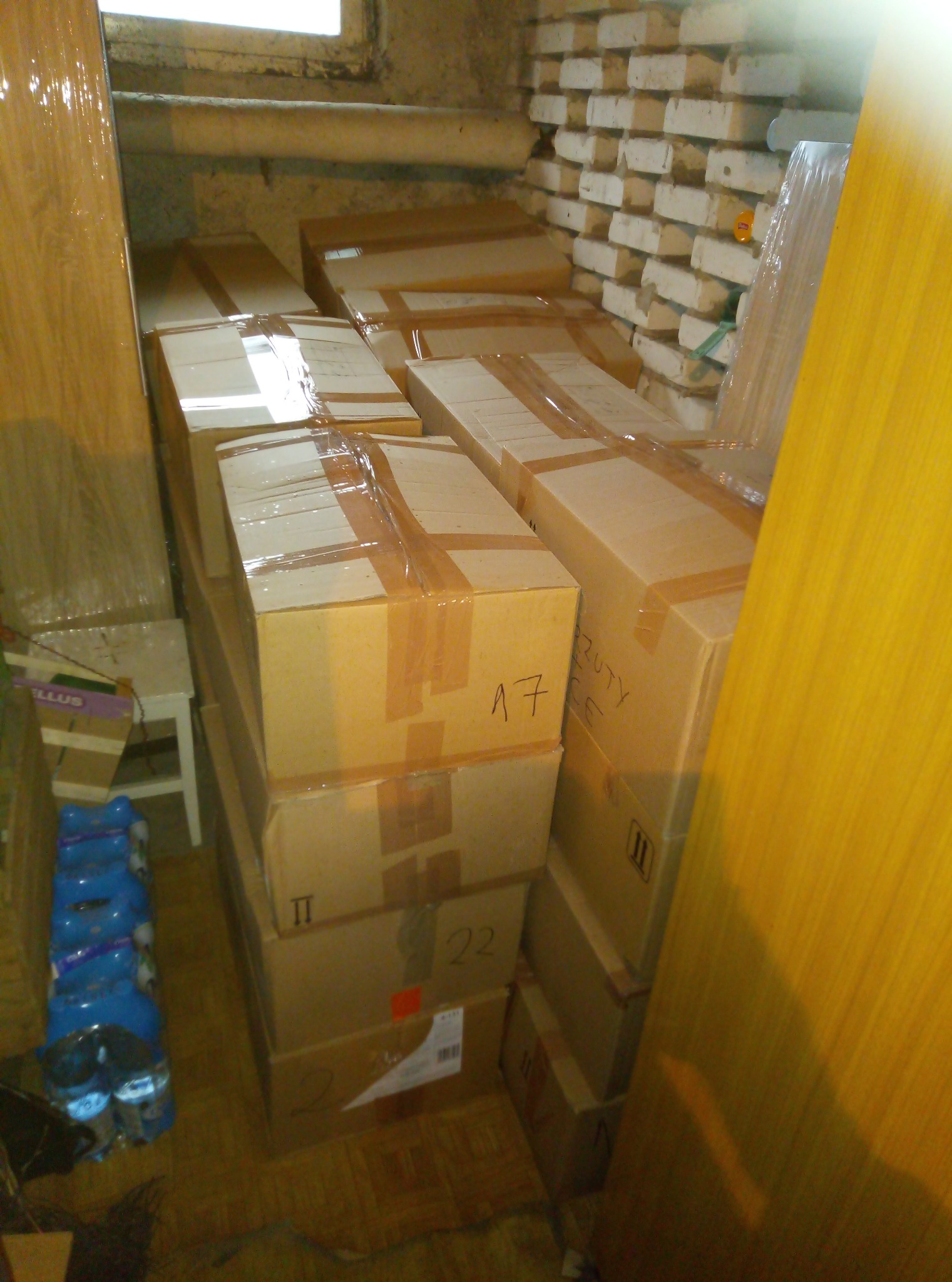 Stos kartonowych pudeł zabezpieczonych taśmą, częściowo opisanych markerem, ustawionych w piwnicy z ceglaną ścianą i drewnianą podłogą, obok plastikowych butelek z wodą.