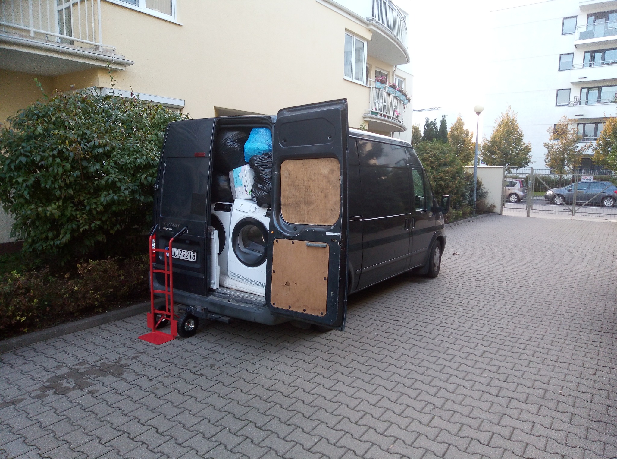 Czarny van z otwartymi drzwiami bagażnika, załadowany pralką, materacem i workami z rzeczami, przed budynkiem mieszkalnym; czerwony wózek transportowy z tyłu pojazdu.