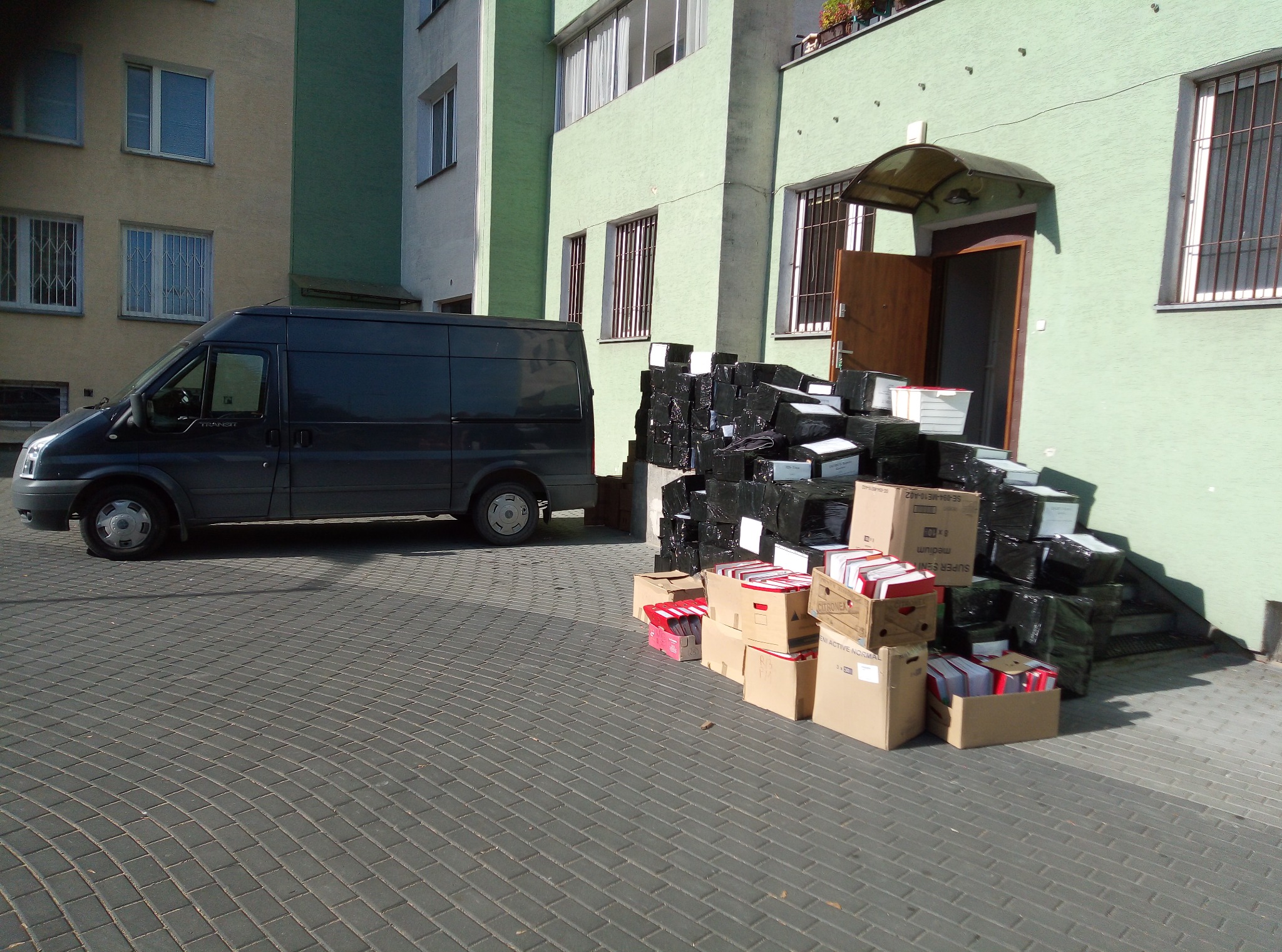 Czarny van dostawczy zaparkowany obok sterty zapakowanych kartonów i folii przed wejściem do budynku.