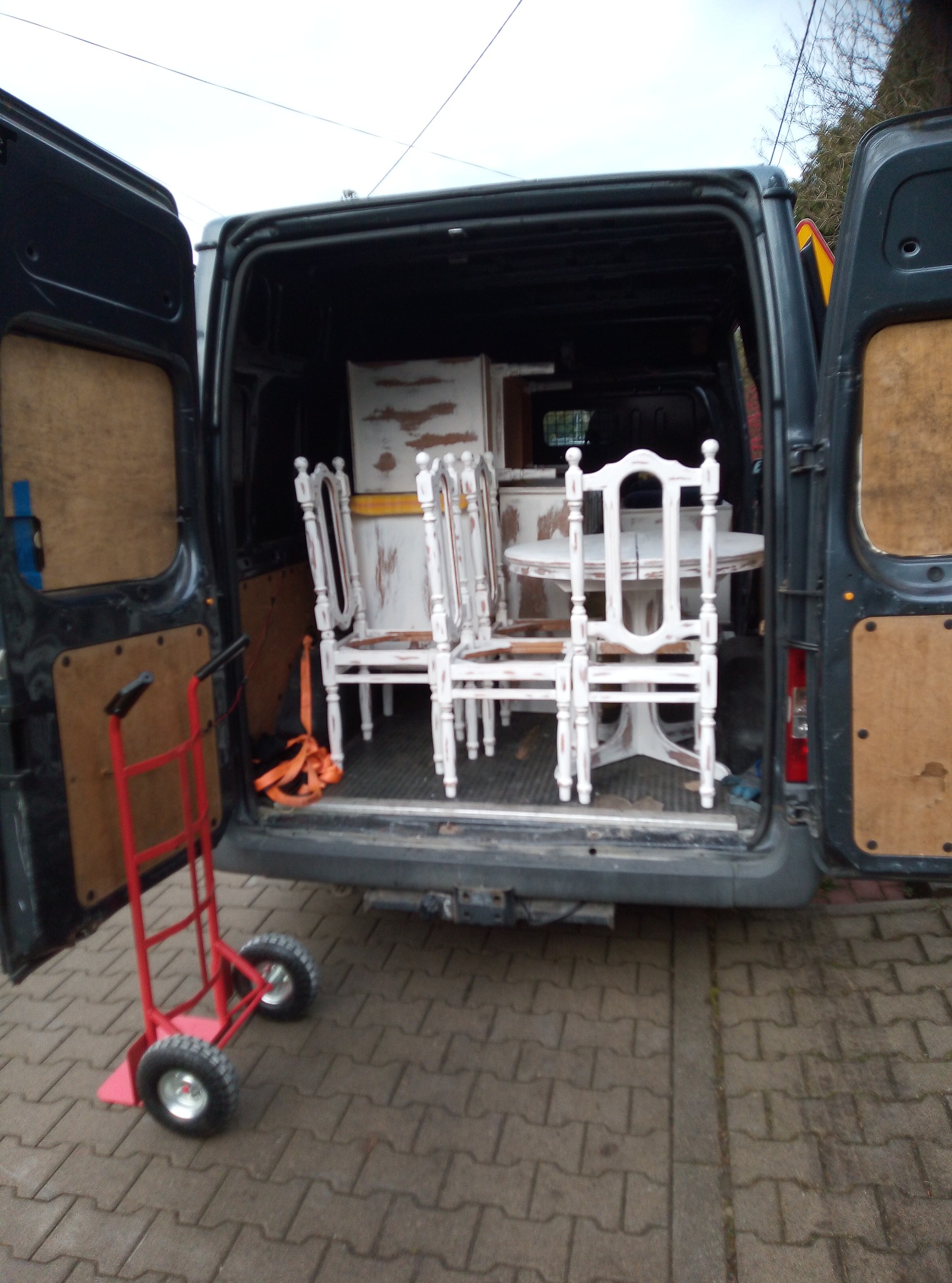 Wnętrze busa z otwartymi drzwiami, wypełnione białymi, postarzanymi meblami w stylu vintage: stół i krzesła. Czerwony wózek transportowy stoi obok, gotowy do rozładunku.