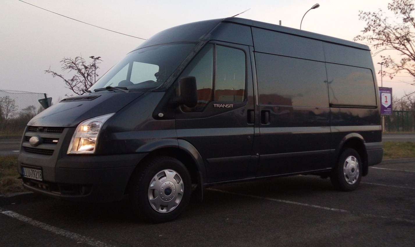 Ciemnoszary Ford Transit z podwyższonym dachem, zaparkowany na parkingu, z widoczną tablicą rejestracyjną i odbiciem w szybach.