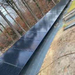 Długi rząd paneli fotowoltaicznych zainstalowanych na zboczu, zabezpieczonym czarną geowłókniną, w tle widoczne drzewa.