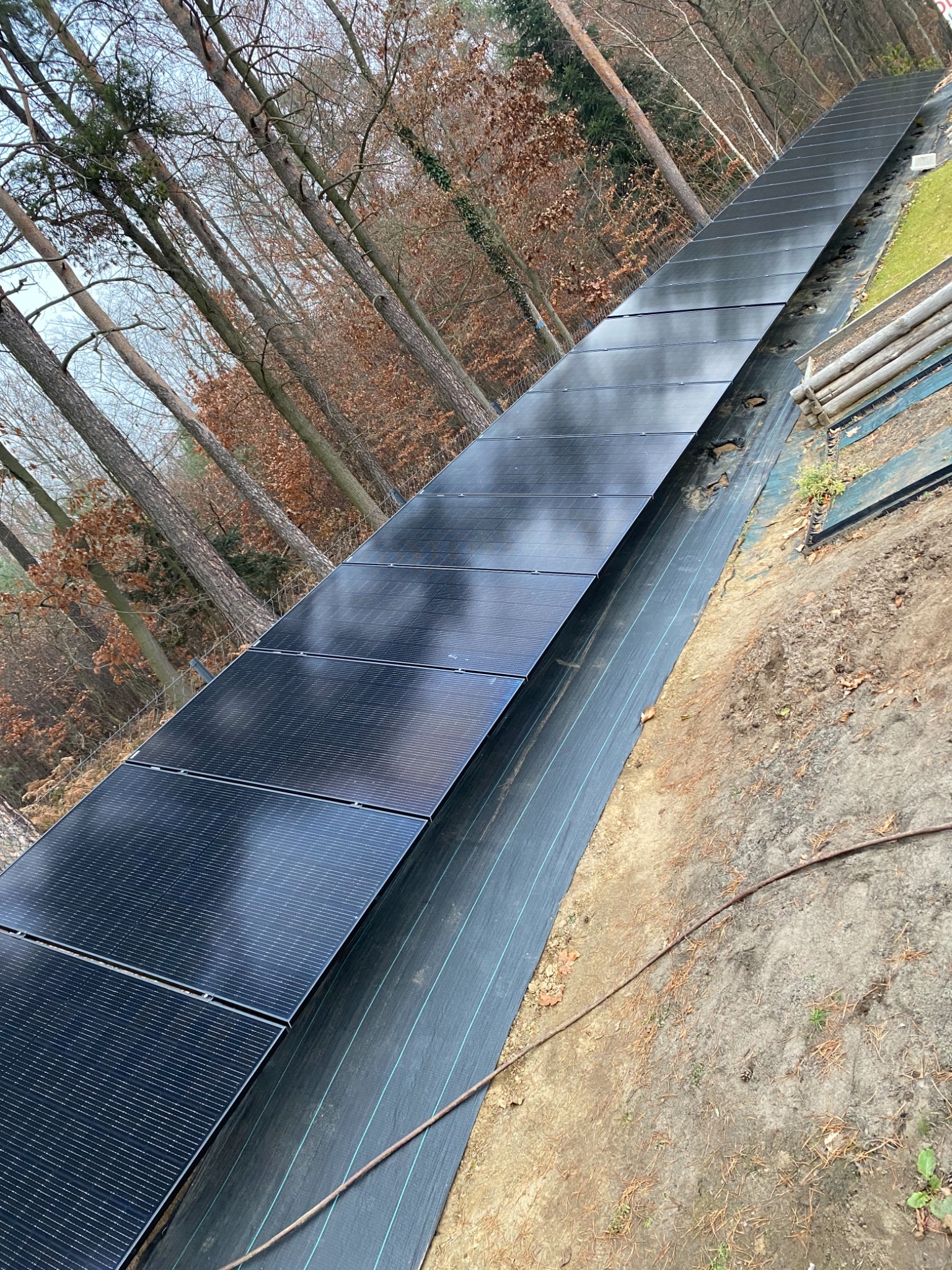 Długi rząd paneli fotowoltaicznych zainstalowanych na zboczu, zabezpieczonym czarną geowłókniną, w tle widoczne drzewa.