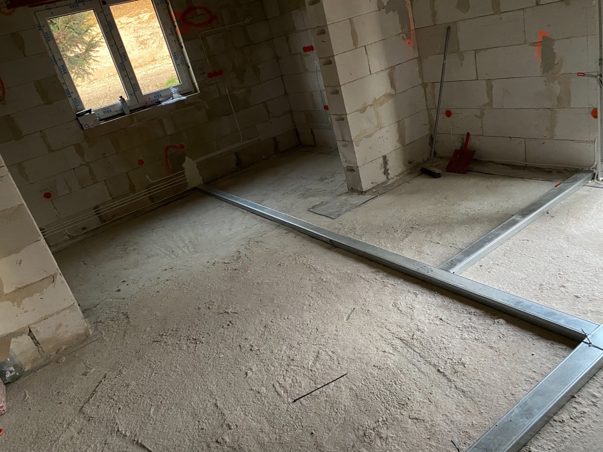 Surowy stan wnętrza z instalacją elektryczną: widoczne przewody w ścianach z betonu komórkowego, okno z widokiem na drzewo, metalowa konstrukcja pod podłogę, ślady po wyznaczaniu punktów instalacji.