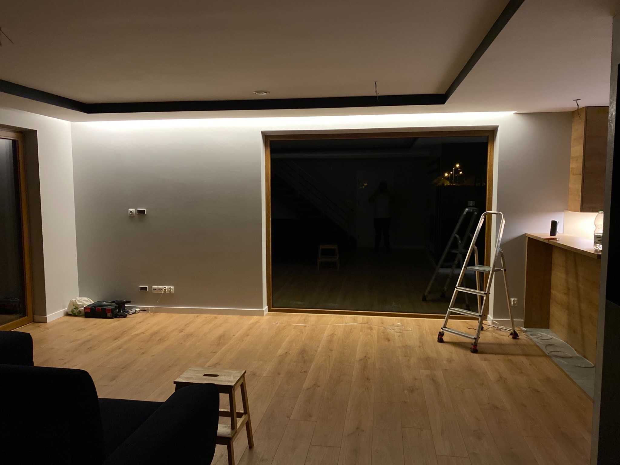 Wnętrze pokoju z zamontowanym oświetleniem LED w suficie, drabina oparta o ścianę, widoczne narzędzia i skrzynka narzędziowa na podłodze, przewody wystające z sufitu.