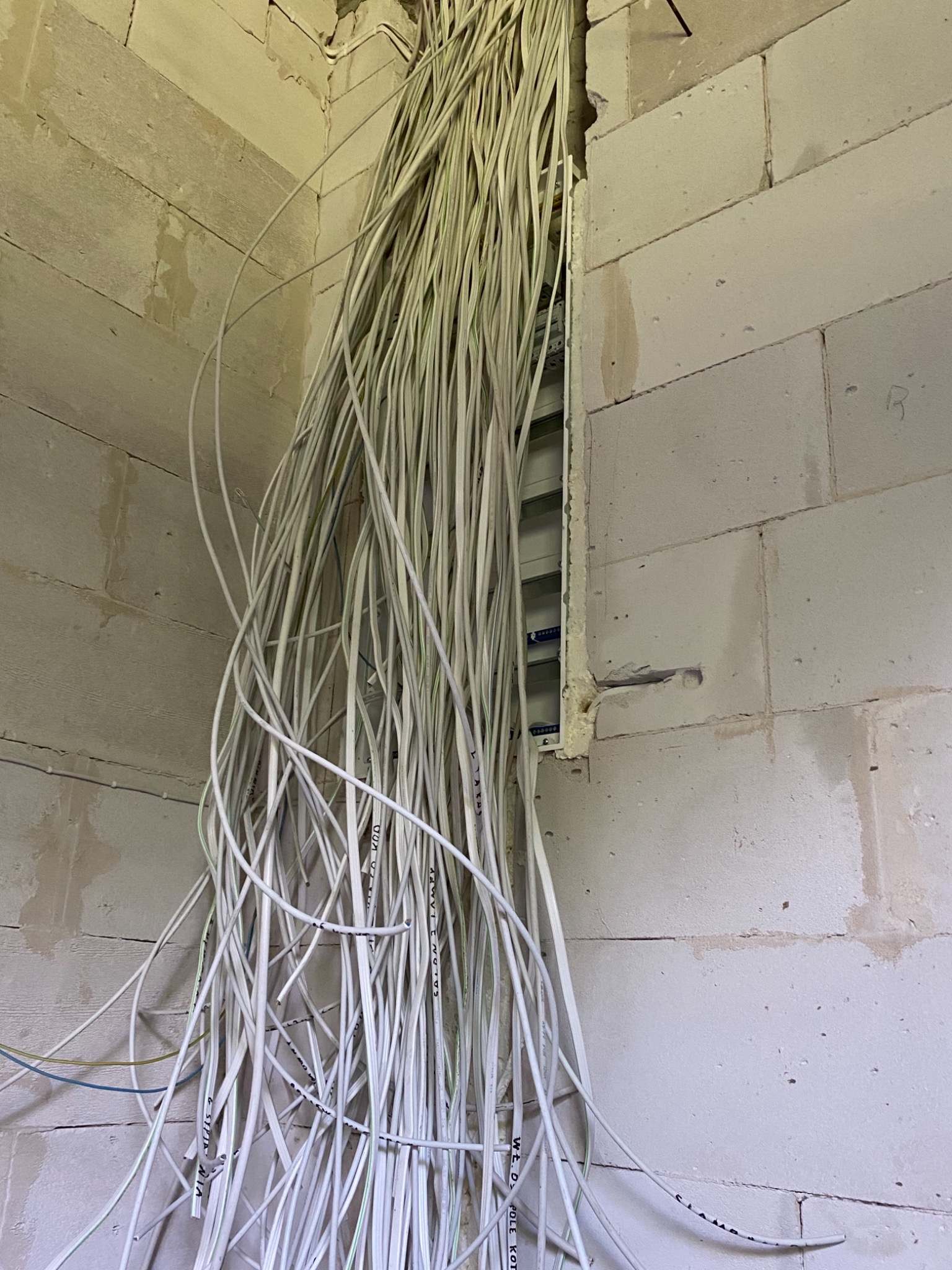 Wiązka oznaczonych przewodów zwisających ze ściany z bloków betonu komórkowego, w tle widoczna skrzynka rozdzielcza w przygotowanym otworze.
