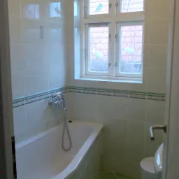 Jasna łazienka z wanną, umywalką i oknem. Ściany wyłożone jasnymi płytkami z dekoracyjnym paskiem, podłoga w zielonych płytkach. Widok zza otwartych drzwi.