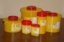 Żółte pojemniki z czerwonymi pokrywami, oznaczone symbolem biohazard, ustawione na brązowym blacie. Różne wielkości pojemników na odpady medyczne.