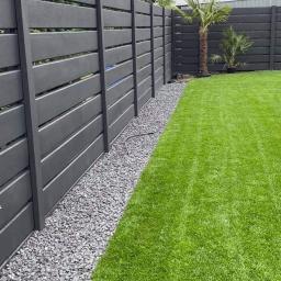 BrukiOgród Maciej Świątek - Nowoczesne ogrodzenie panelowe w kolorze grafitowym z poziomymi deskami, oddzielone od trawnika pasem szarych kamieni, z palmą i zielenią w tle.