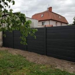 BrukiOgród Maciej Świątek - Ciemnoszary płot z poziomych desek, widok z zewnątrz posesji na tle domu z czerwonym dachem i zielenią drzew.