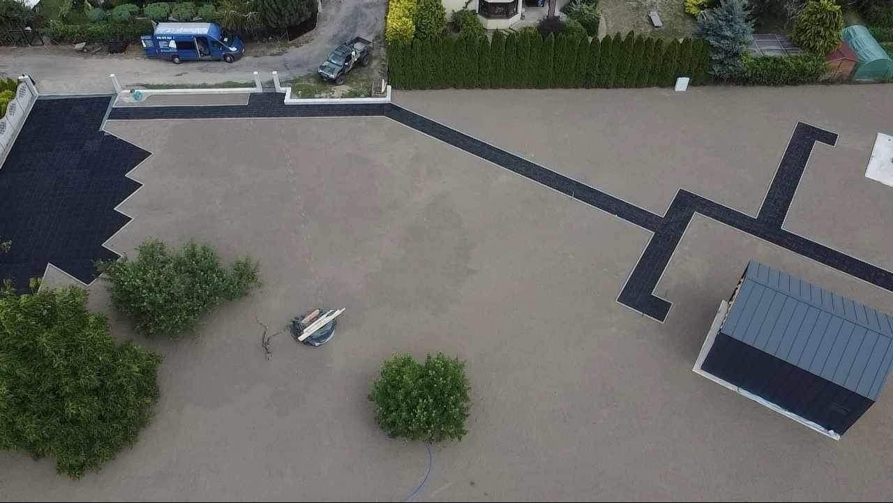 Widok z drona na posesję z częściowo ułożoną, ciemną kostką brukową w geometryczne wzory, widoczne narzędzia i samochody w tle. Etap prac brukarskich, zieleń wokół.