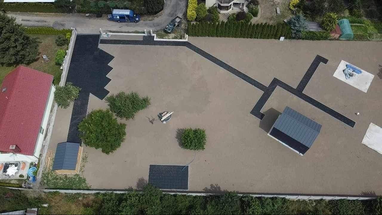 Widok z góry na posesję z ułożoną kostką brukową w geometryczne wzory, ciemne ścieżki przecinają jasną nawierzchnię, w tle dom z czerwonym dachem i zieleń.