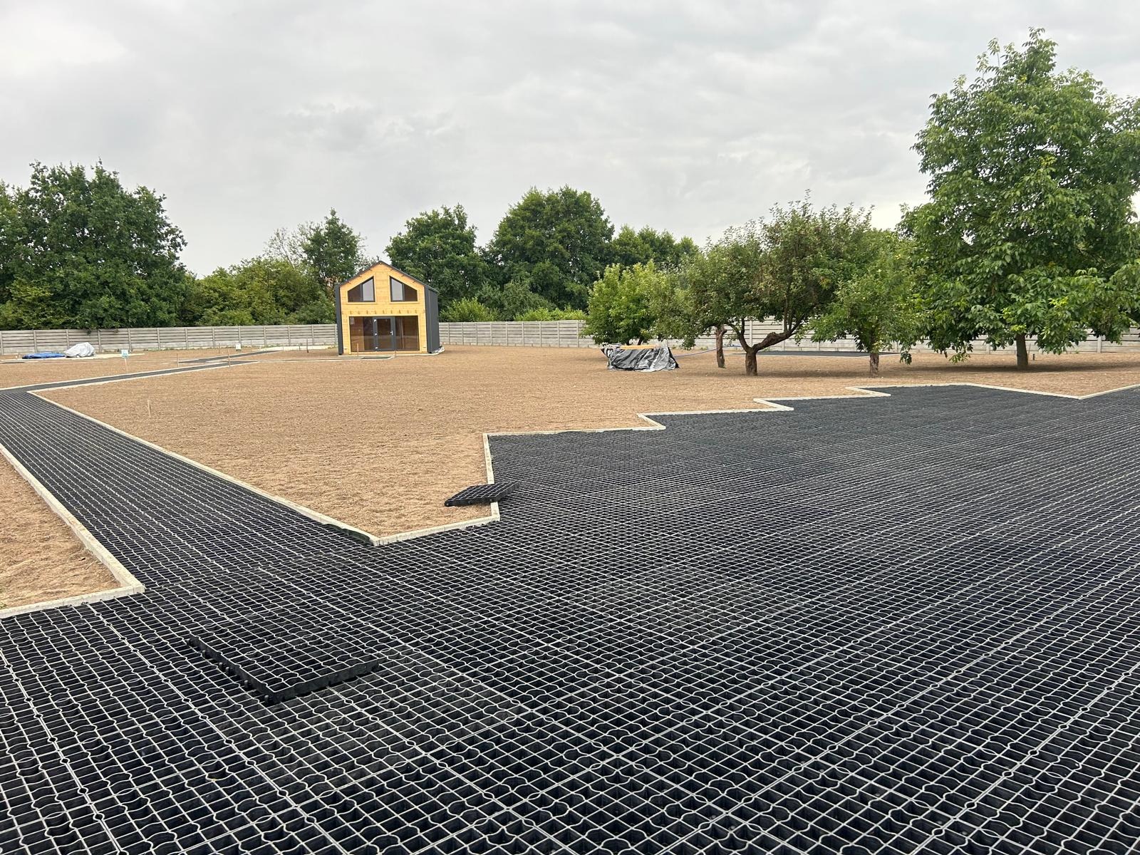 Układanie geokraty na piasku w ogrodzie, widoczny domek narzędziowy i ogrodzenie panelowe w tle. Częściowo ułożona nawierzchnia, etap prac.