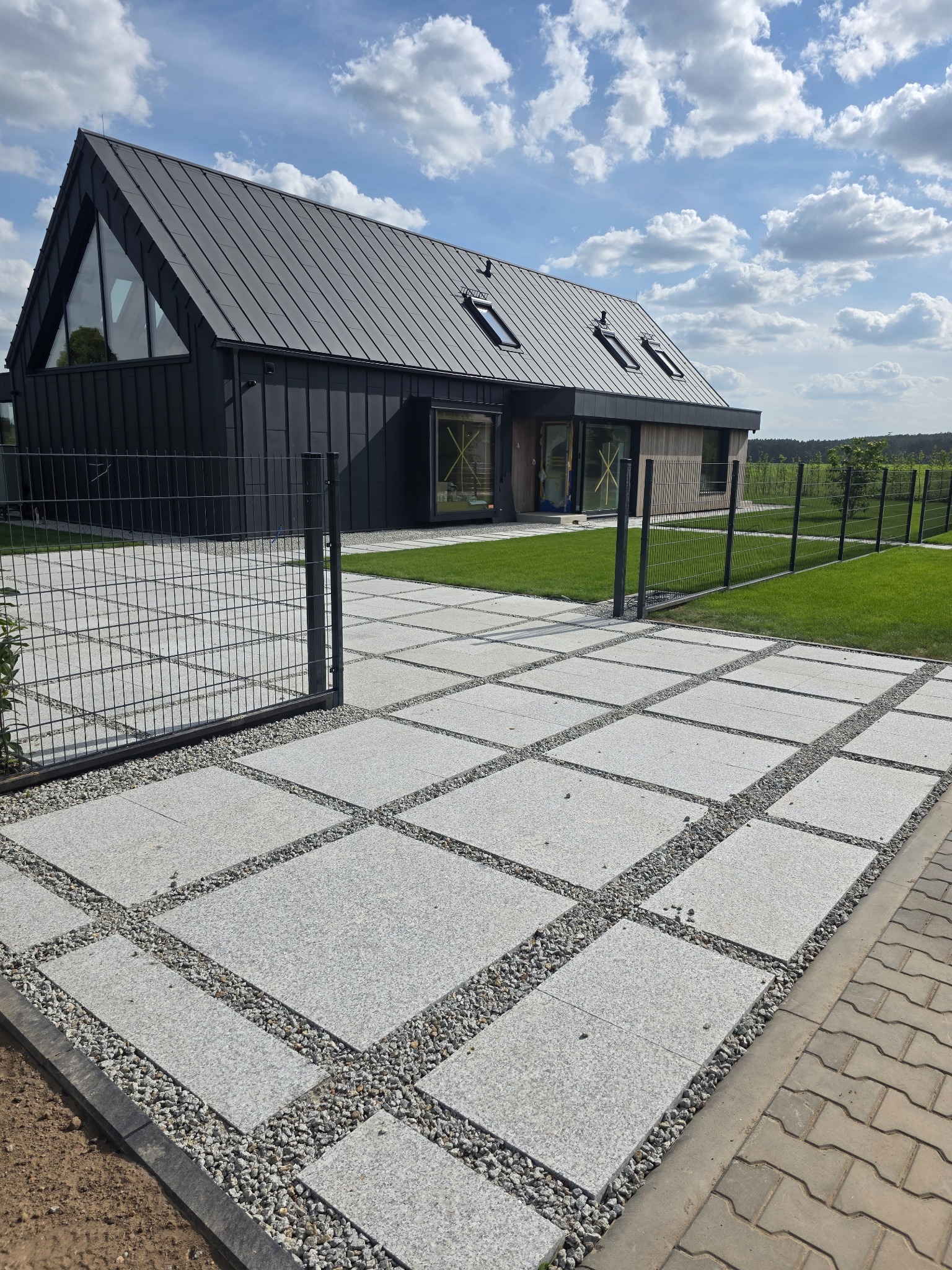 Nowoczesny dom z szarej kostki brukowej, ogrodzony panelowym ogrodzeniem. Widoczne elementy architektury krajobrazu i błękitne niebo.
