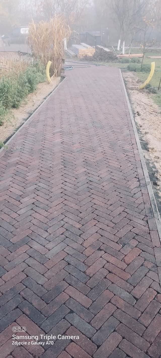 Ułożona kostka brukowa w jodełkę, tworząca ścieżkę w ogrodzie. Widoczne trawy ozdobne i elementy instalacji nawadniającej. Mglista pogoda w tle.