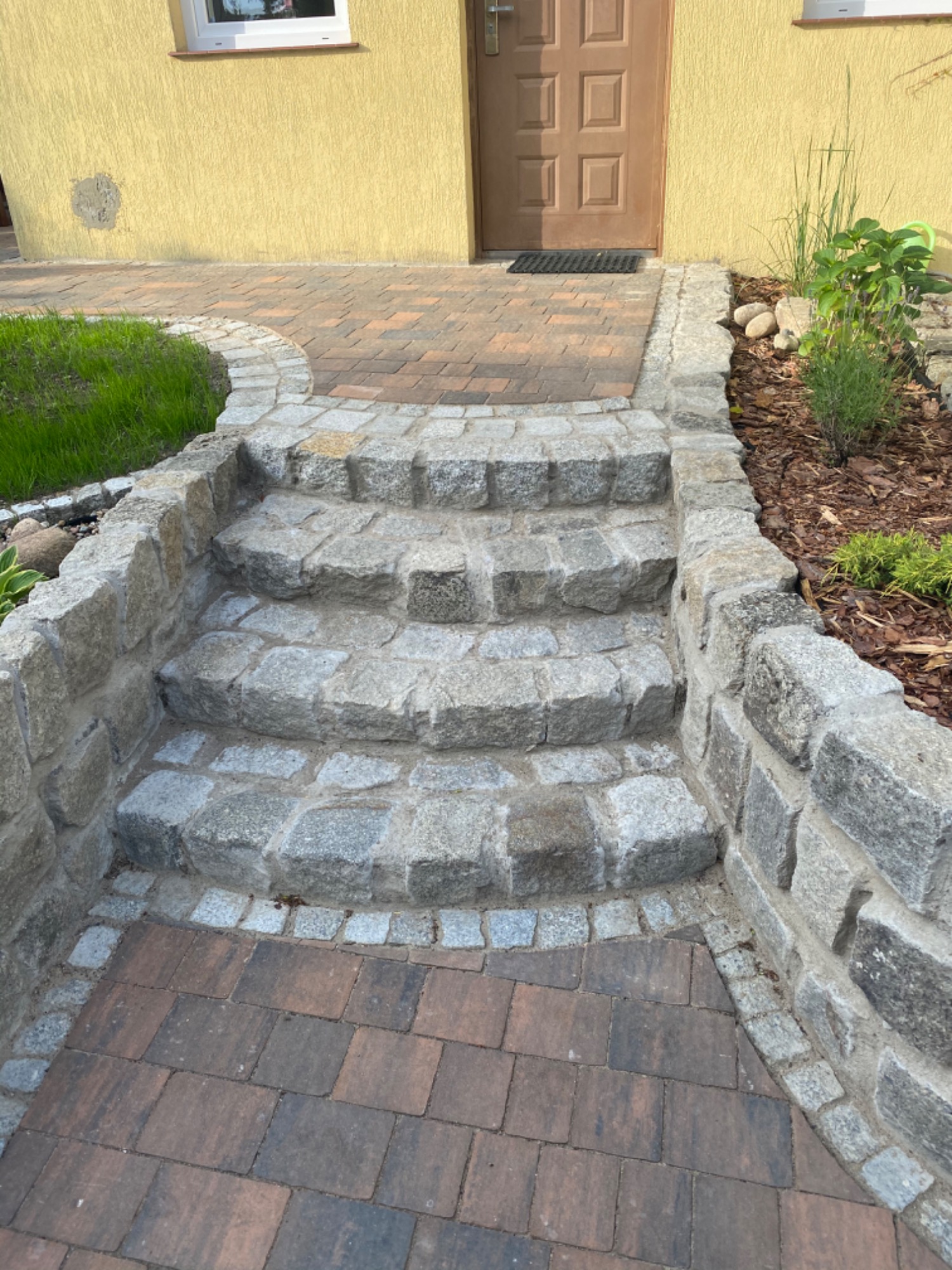 Granitowe schody wejściowe do domu z kostki granitowej, otoczone obrzeżem z kostki, prowadzące do drzwi wejściowych.