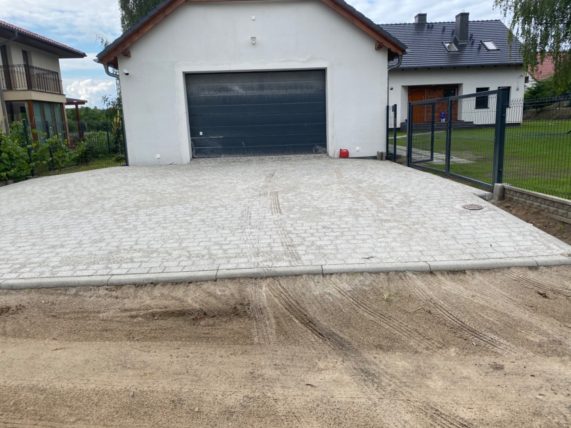 Jasnoszara kostka brukowa ułożona na podjeździe przed garażem i domem jednorodzinnym, widoczne ślady opon na piasku obok kostki, obrzeża betonowe.