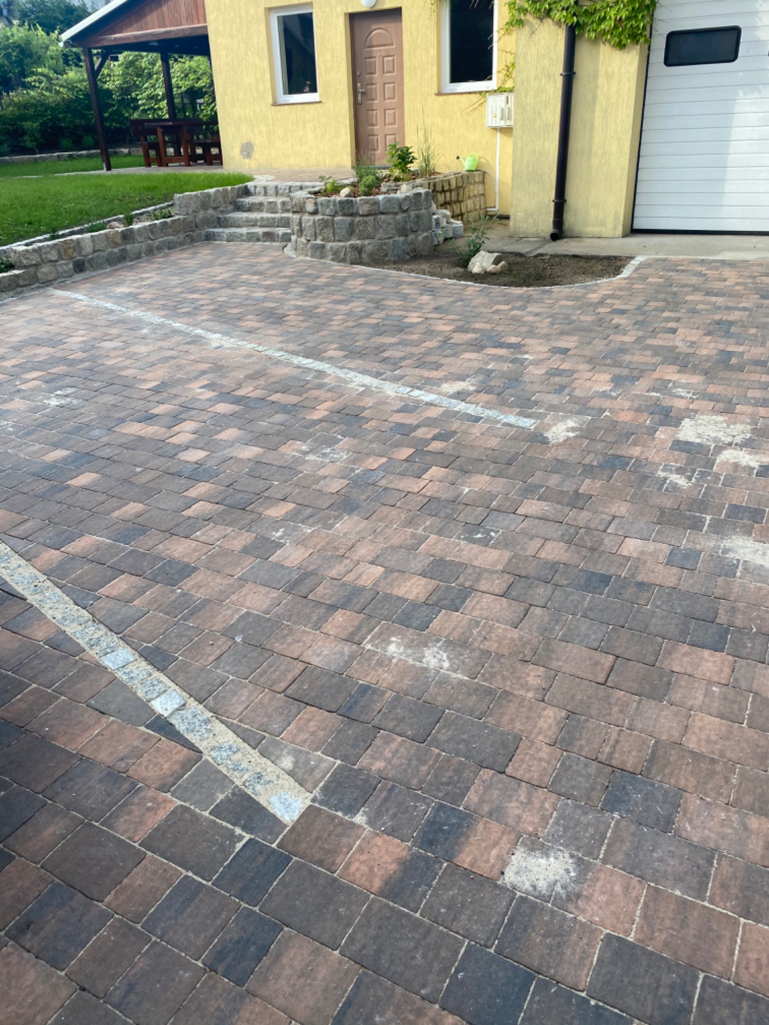 Ułożona kostka brukowa w odcieniach brązu i szarości tworząca podjazd przed domem z żółtą elewacją i schodami z kamienia.