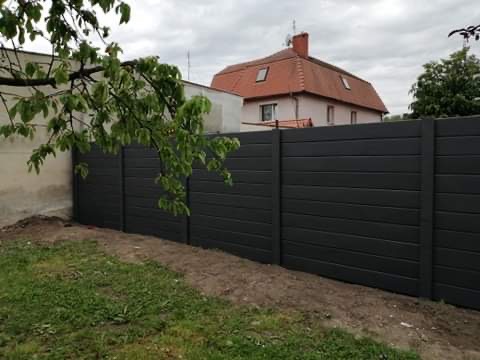 Ciemnoszary płot z poziomych desek, widok z zewnątrz posesji na tle domu z czerwonym dachem i zielenią drzew.