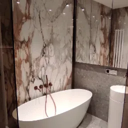 Nowoczesna łazienka z wolnostojącą białą wanną, marmurową ścianą i miedzianą armaturą, oświetlona punktowo.