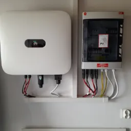 Dwa białe panele elektryczne, jeden marki Huawei z zielonymi diodami, zamontowane na białej ścianie nad tablicą z zapisanymi równaniami.