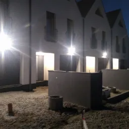 Nowo wybudowane szeregówki z zamontowanym oświetleniem zewnętrznym, widoczne niedokończone prace ziemne i zaparkowany samochód.