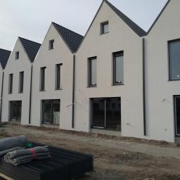 A.C. Instal - Rząd nowych, identycznych domów z białymi ścianami i czarnymi oknami, jeszcze w trakcie budowy, z widocznymi kablami wystającymi ze ścian i materiałami budowlanymi leżącymi na ziemi.