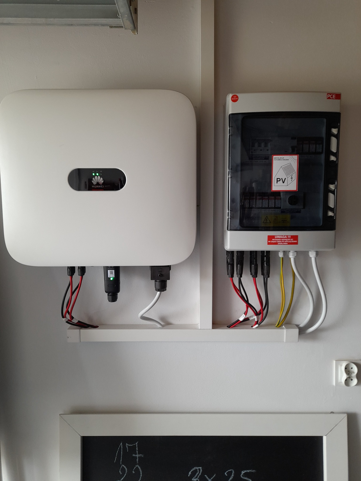 Dwa białe panele elektryczne, jeden marki Huawei z zielonymi diodami, zamontowane na białej ścianie nad tablicą z zapisanymi równaniami.