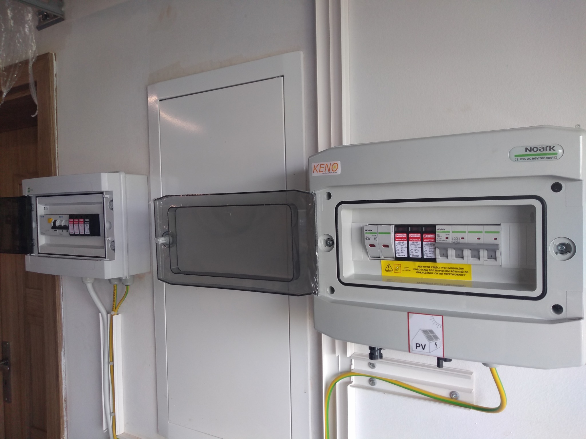Rozdzielnice elektryczne, w tym jedna z oznaczeniem PV, zamontowane na ścianie, z widocznymi przewodami ochronnymi i elementami zabezpieczeń. Jedna rozdzielnica otwarta, ukazująca wnętrze.
