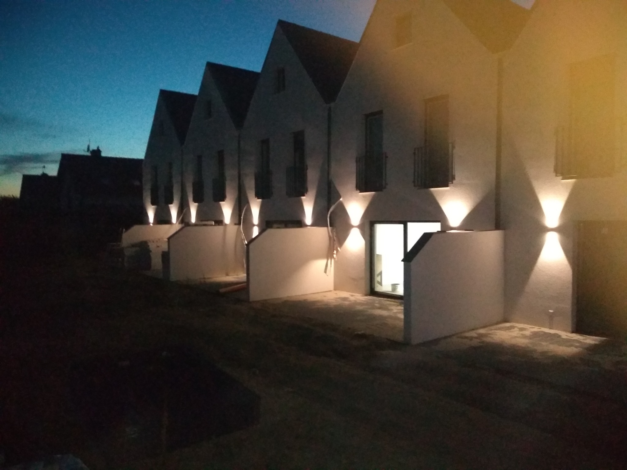 Nowoczesne segmenty domów jednorodzinnych oświetlone zewnętrznymi lampami ściennymi, widok nocny z nierównym terenem w tle i elementami budowlanymi.