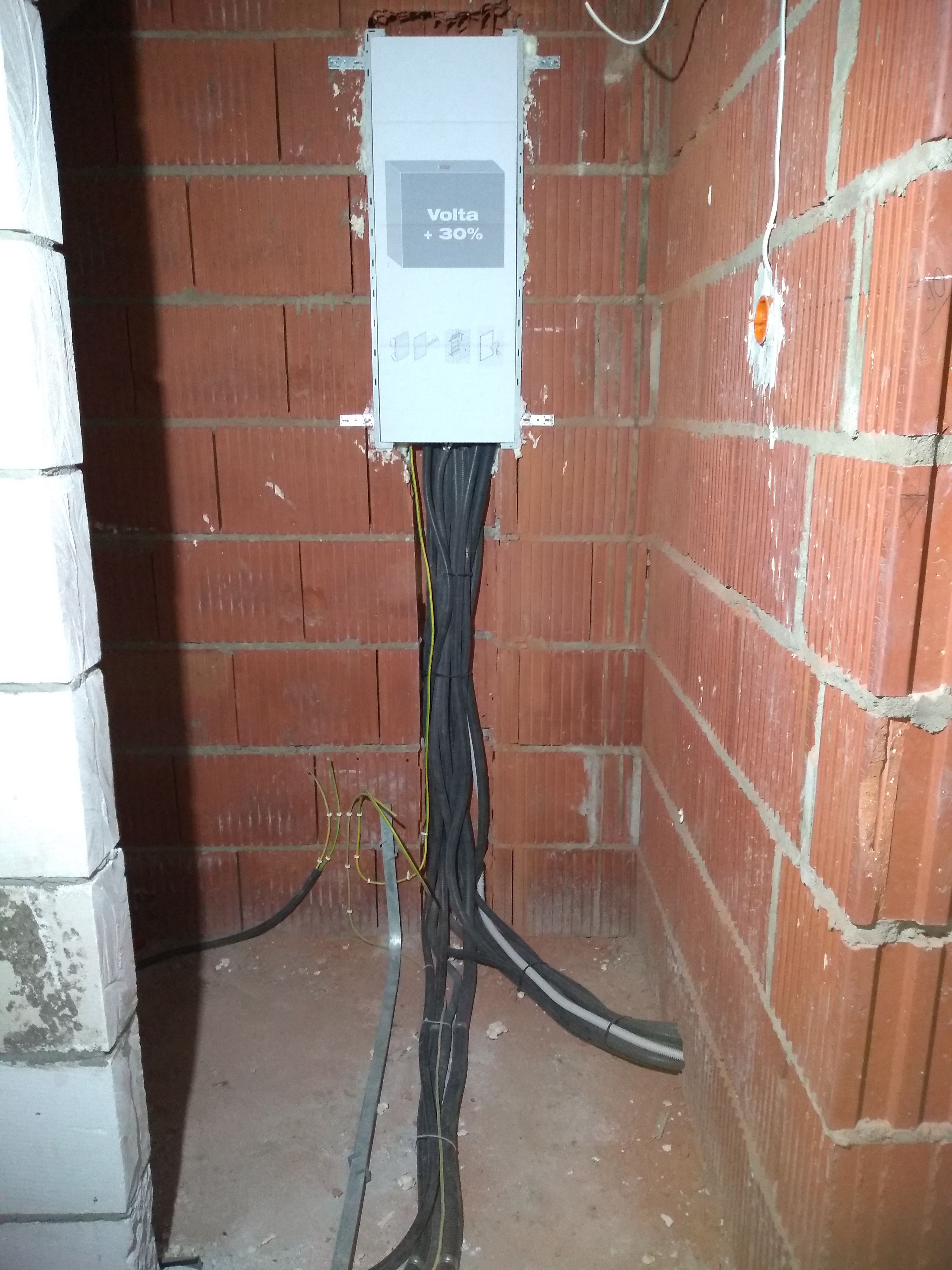 Szara skrzynka elektryczna Volta z podłączonymi kablami w rurach karbowanych, zamontowana na ścianie z czerwonej cegły dziurawki w surowym stanie, z widocznym przewodem uziemiającym i puszką...
