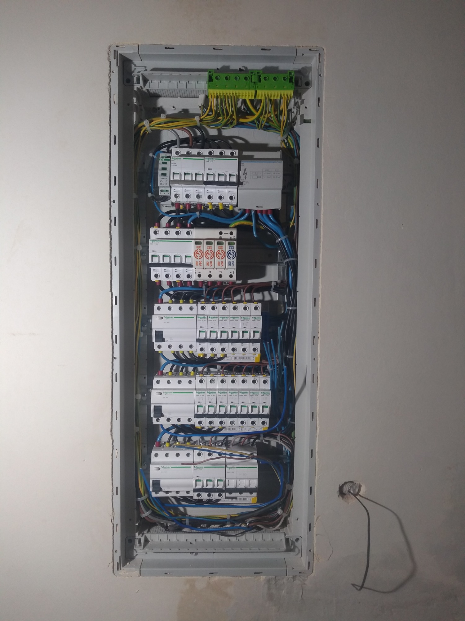 Rozdzielnica elektryczna z zainstalowanymi bezpiecznikami i okablowaniem, widoczna wnęka w ścianie z wystającym przewodem elektrycznym.