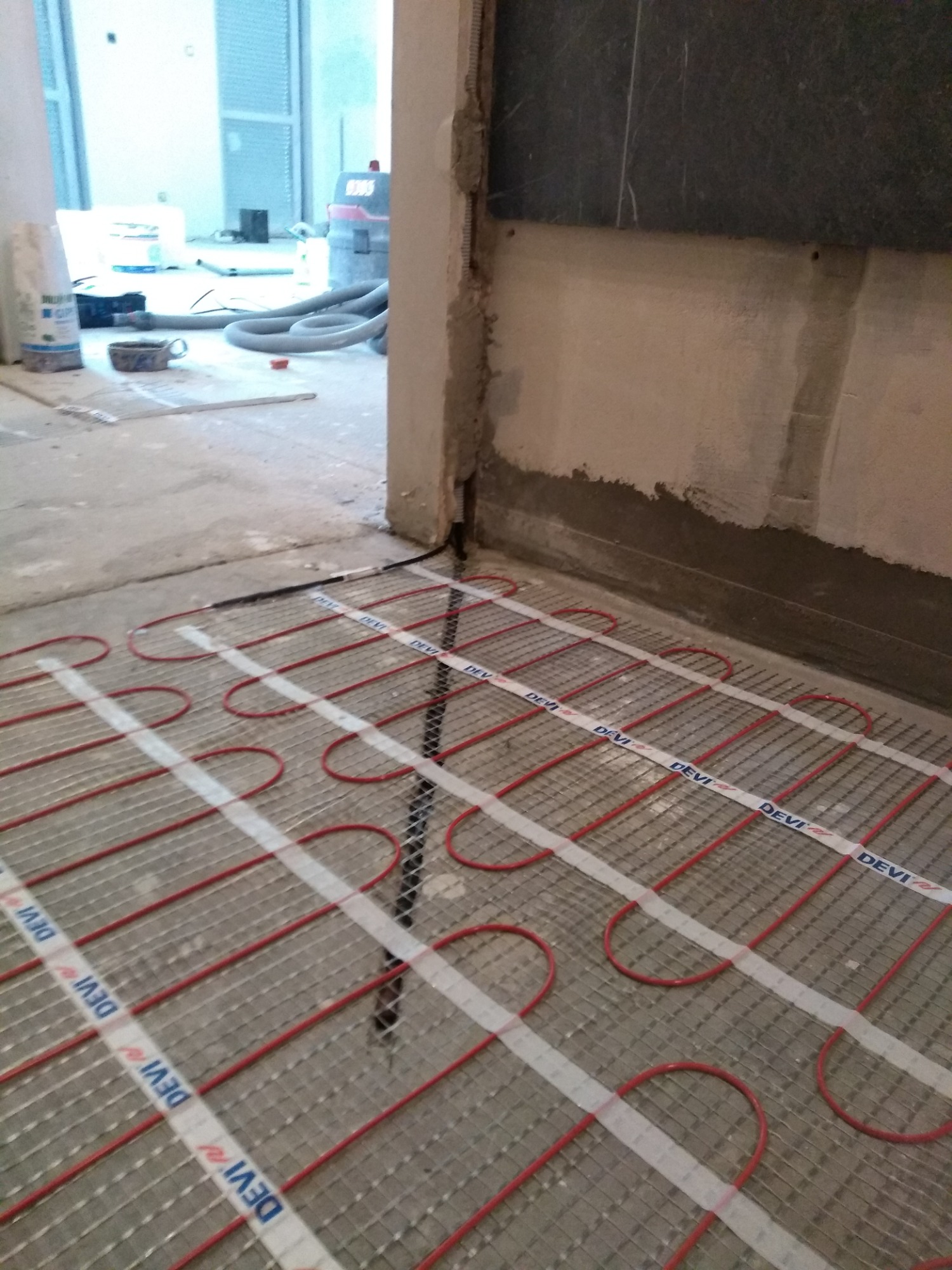 Rozłożona mata grzewcza DEVI na podłodze w trakcie remontu, widoczna instalacja elektryczna pod ogrzewanie podłogowe, surowy beton i fragment ściany w tle.