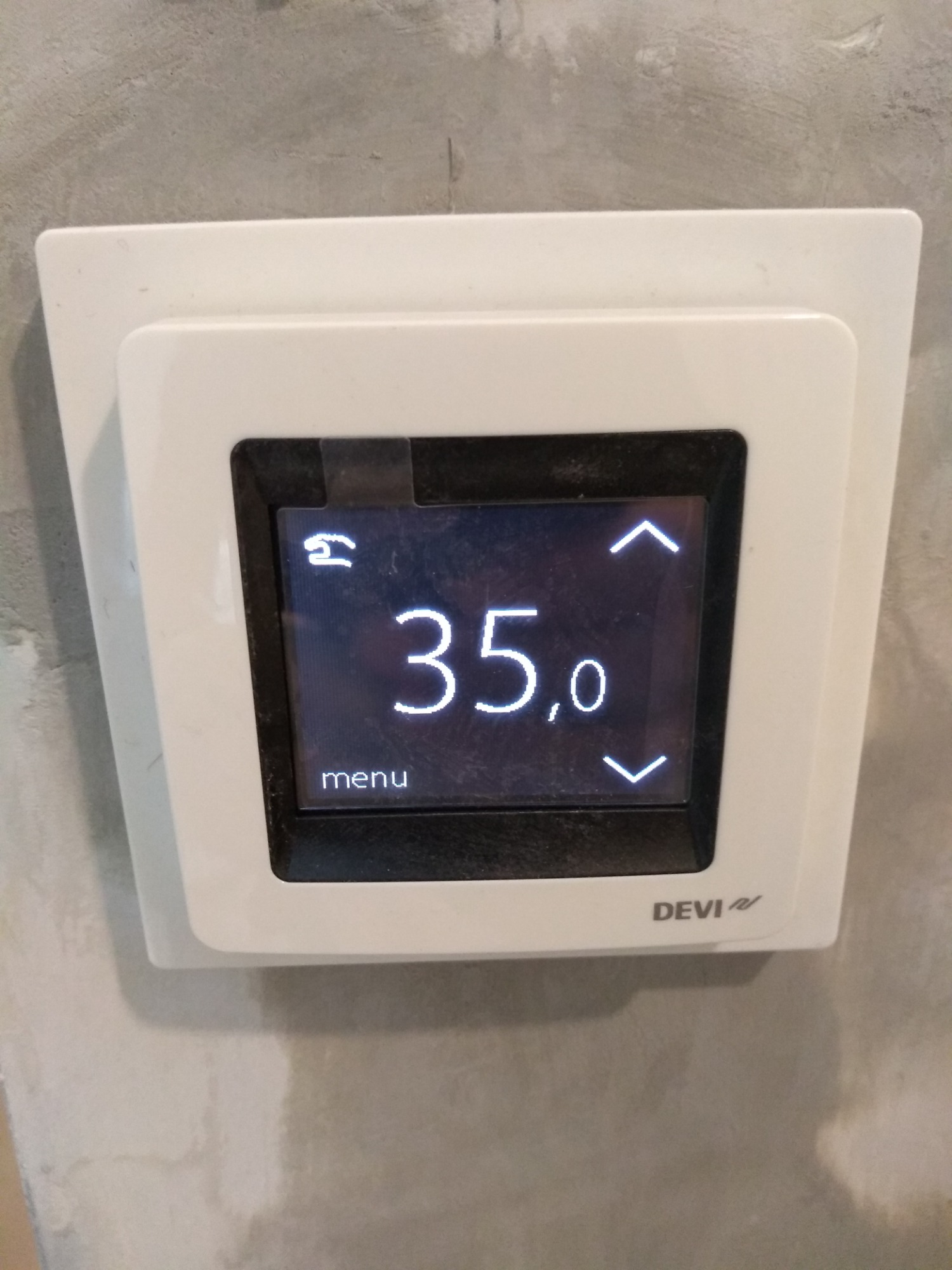 Biały termostat Devi zamontowany na szarej, surowej ścianie, wyświetlający temperaturę 35 stopni Celsjusza.