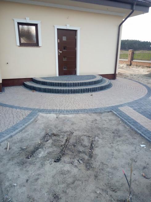 Fragment układania kostki brukowej przed domem z widocznymi dwustopniowymi schodami i przygotowanym wykopem pod dalszą część nawierzchni.
