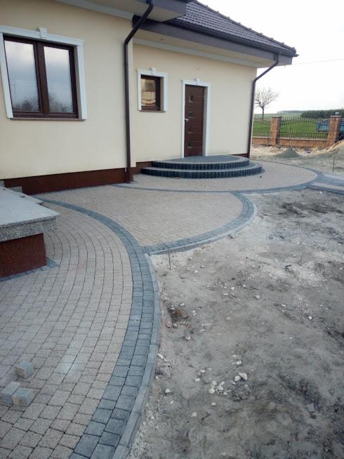 Wykończony podjazd z beżowej kostki brukowej z szarym obramowaniem prowadzący do schodów wejściowych domu, widoczne niezakończone prace ziemne obok.
